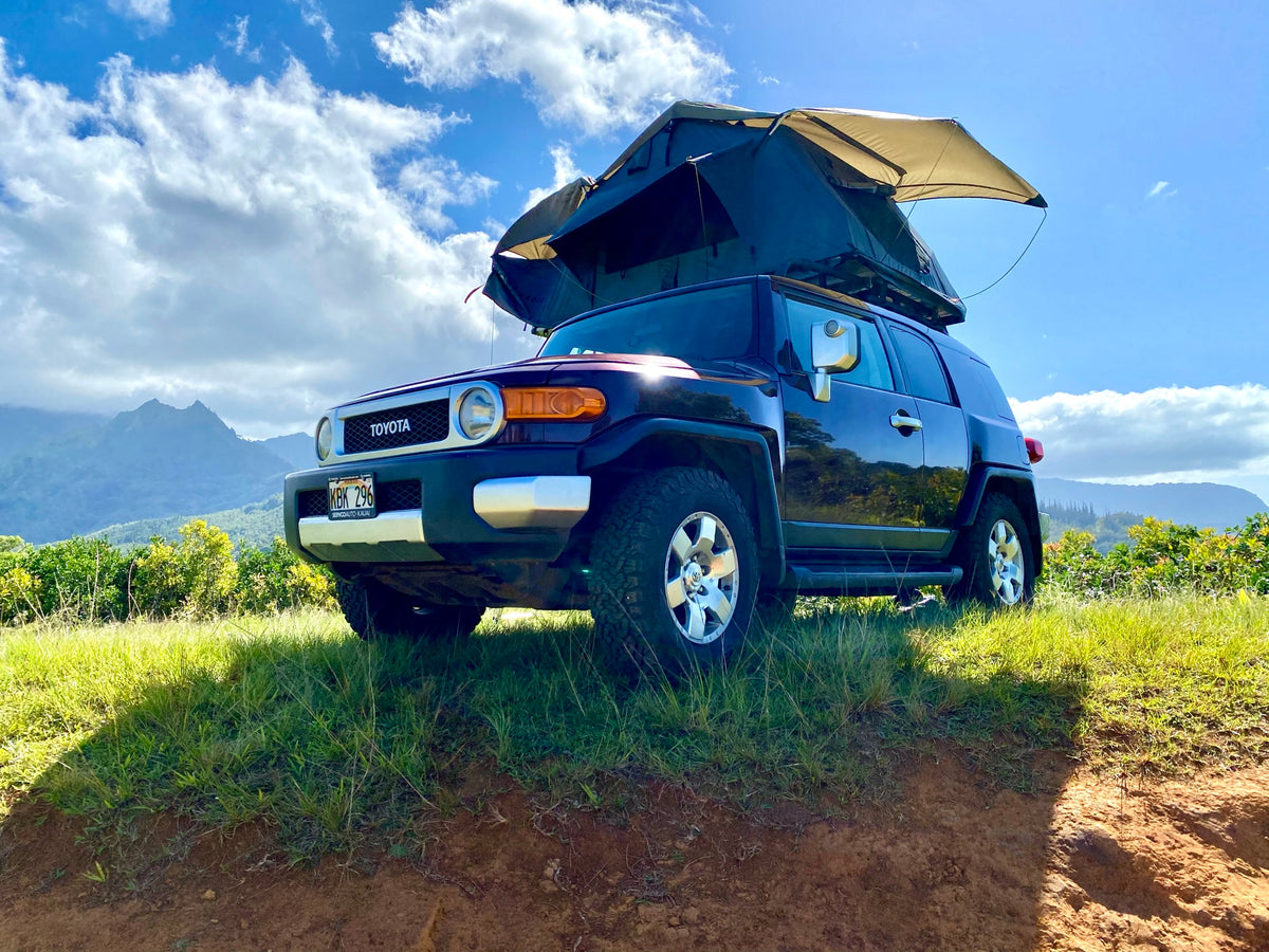 RV rental ready: Dark Toyota FJ Cruiser with open rooftop tent on a sunny, grassy Hawaii hill for outdoor adventure. — photo 10