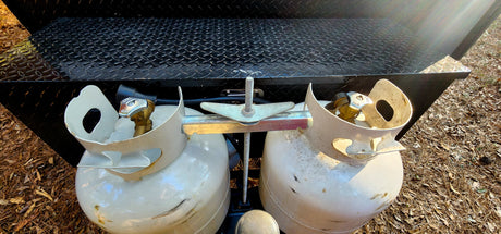 Two white propane tanks are mounted on a black diamond plate surface, typical for an RV rental, with pine needles on the ground.