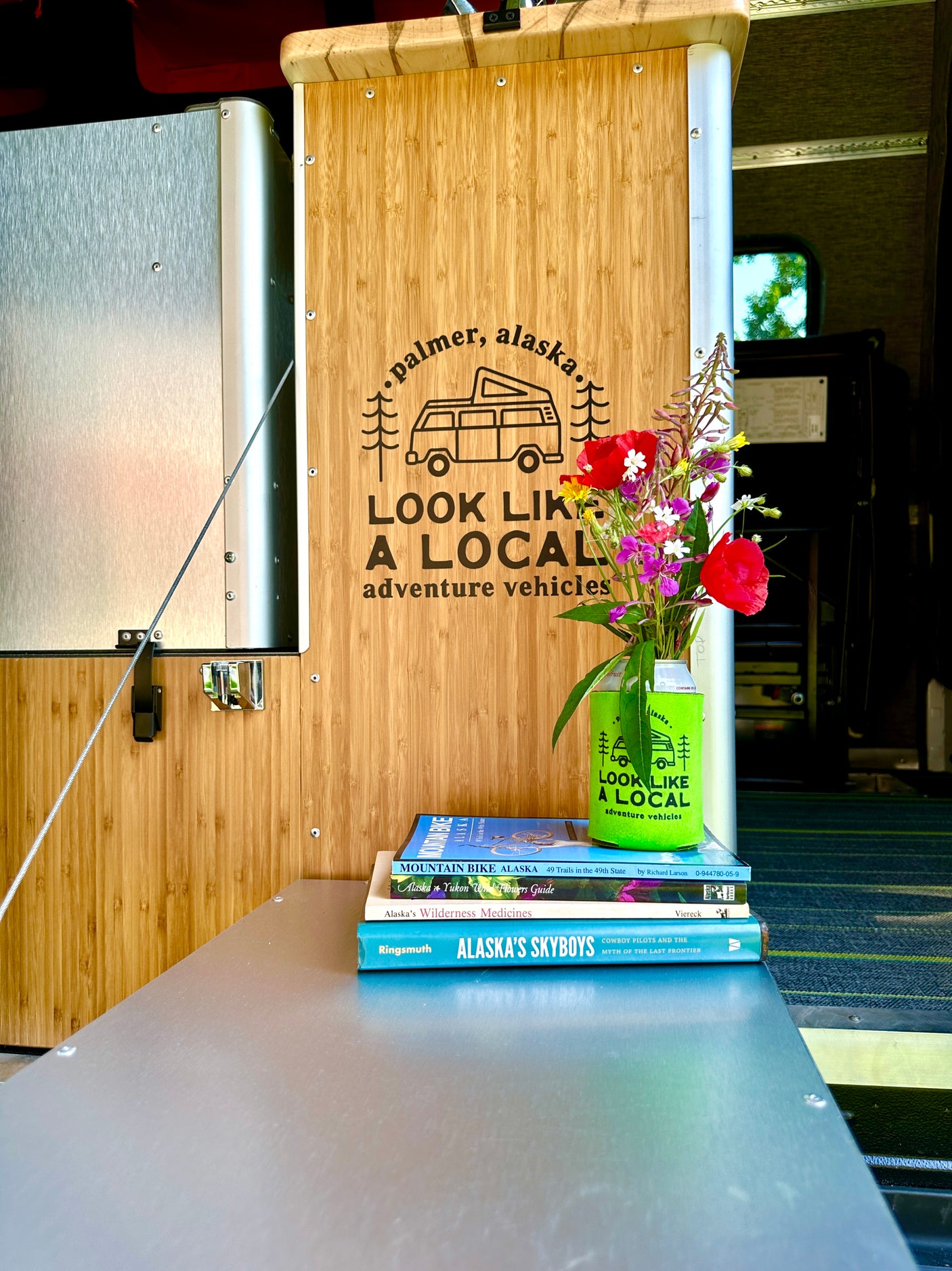 Cozy RV rental interior with custom bamboo panels, "Look Like a Local" branding, Alaska books, and wildflowers.
