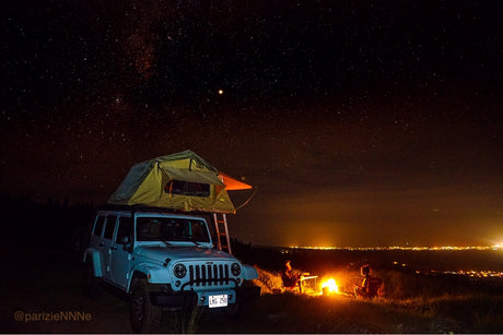 A light blue Jeep with a rooftop tent, offering an RV rental feel, under a starry sky. Two by a campfire overlook city lights at night.