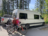 Family with mountain bikes and a white camper van, likely an RV rental, plus a pickup, ready for a forest adventure.