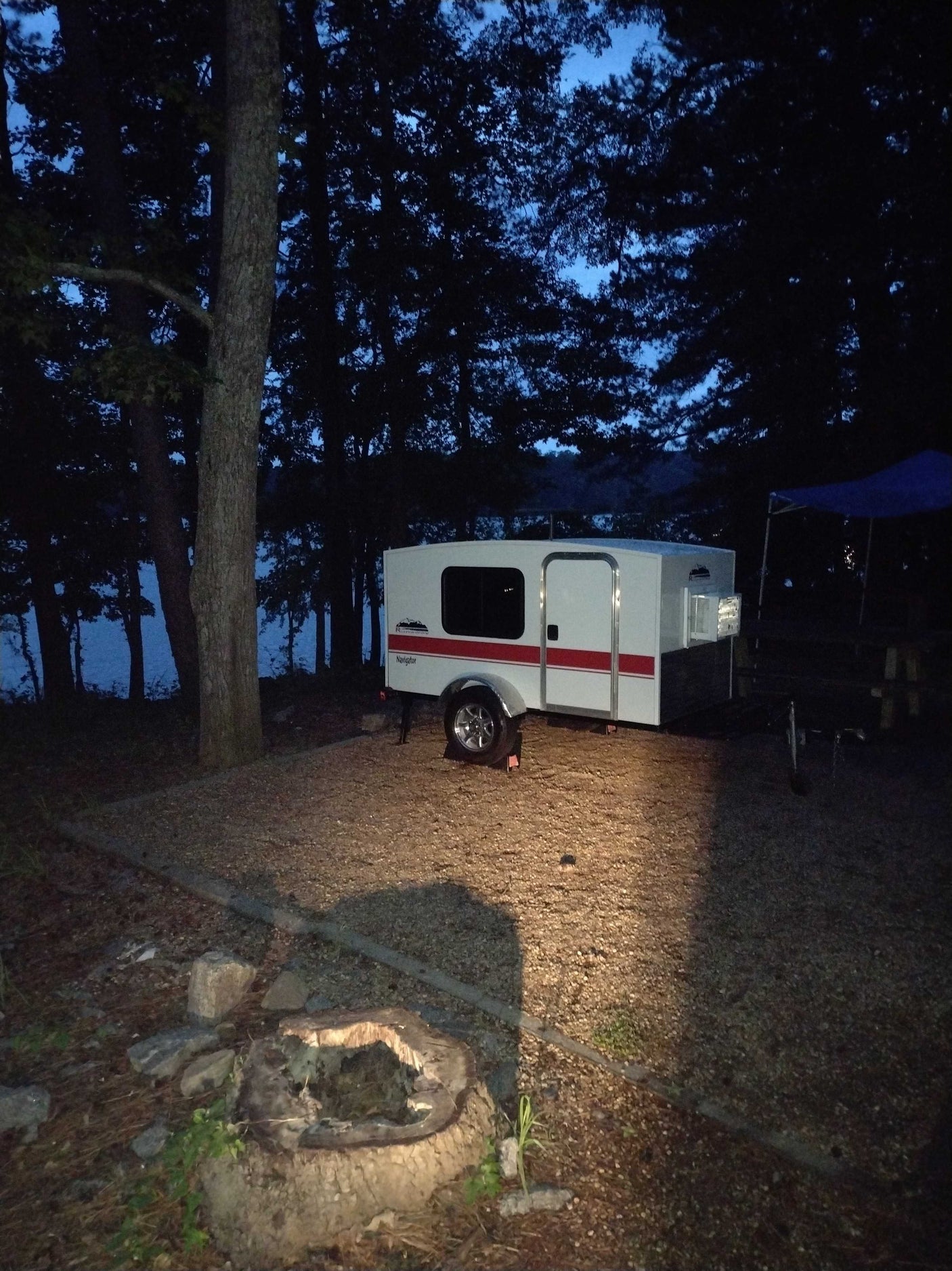 Serene evening campsite: a white rv rental teardrop camper sits by a dark lake, with a fire pit foreground.