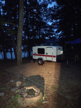 Serene evening campsite: a white rv rental teardrop camper sits by a dark lake, with a fire pit foreground.