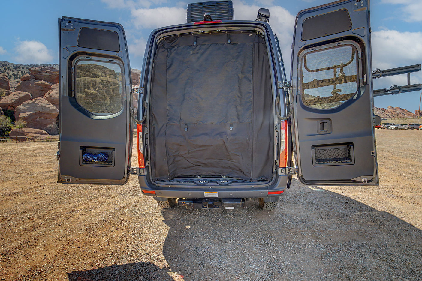 RV rental camper van's open rear doors reveal an interior privacy screen, storage, and bike rack. Parked in a rocky landscape.