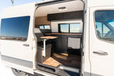View inside an RV rental camper van: modern kitchen with dark cabinets, wood floor, small dining table, and comfortable bed area.
