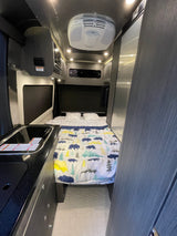 Cozy rv rental interior showing bed with nature-themed comforter, kitchen sink, and overhead AC unit.