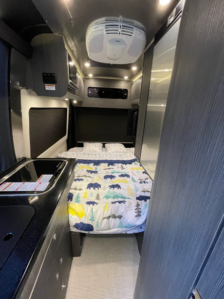 Cozy rv rental interior showing bed with nature-themed comforter, kitchen sink, and overhead AC unit.