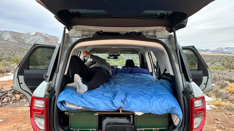 This SUV's cozy car camping setup, a great RV rental alternative, offers mountain views. A person relaxes on the bed with storage below.
