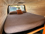 Custom wood paneled RV rental interior with a large comfortable bed and pillows. Rear doors are open, showing the exterior.