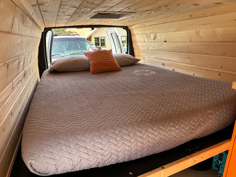 Custom wood paneled RV rental interior with a large comfortable bed and pillows. Rear doors are open, showing the exterior.