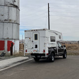 White Northstar truck camper on a black pickup, an ideal RV rental, parked by a metal silo in a rural landscape.