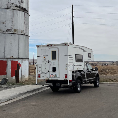 White Northstar truck camper on a black pickup, an ideal RV rental, parked by a metal silo in a rural landscape.