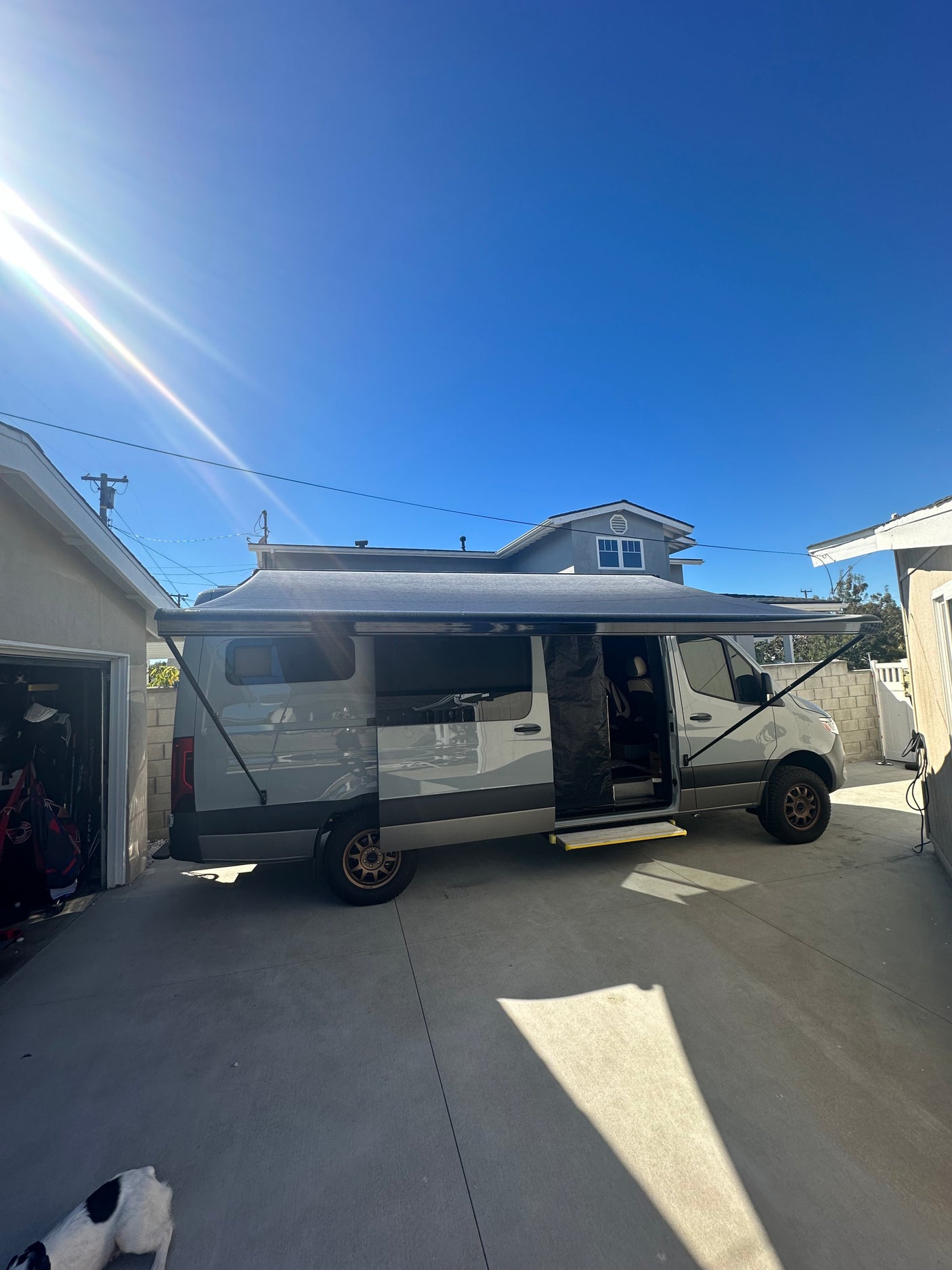 A gray Sprinter camper van, likely an RV rental, with its side awning extended in a bright, sunny driveway.