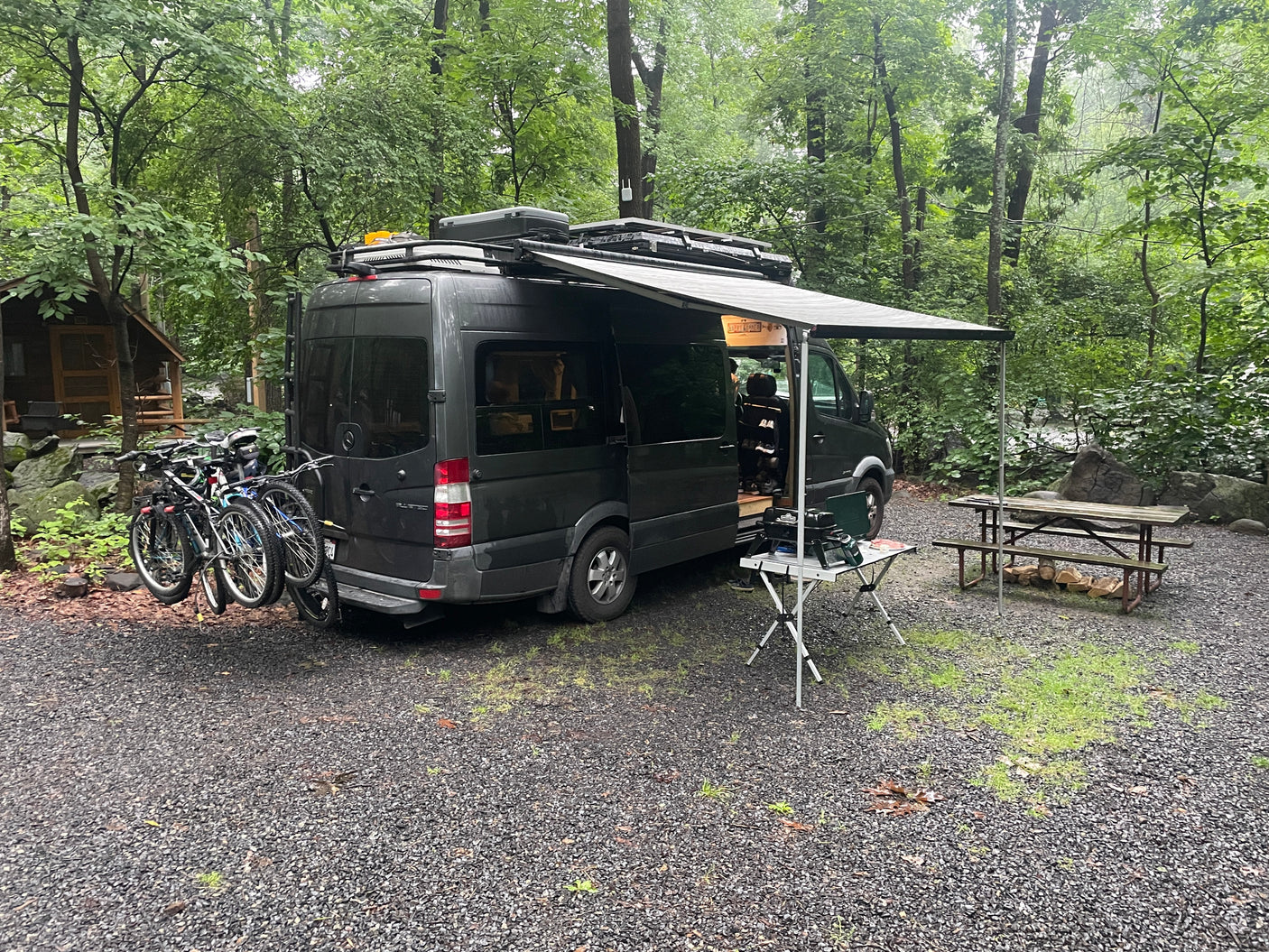 Our RV rental provides ultimate camping comfort! Dark gray van with bikes, awning, & outdoor kitchen setup in nature.