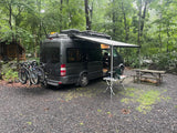 Our RV rental provides ultimate camping comfort! Dark gray van with bikes, awning, & outdoor kitchen setup in nature.