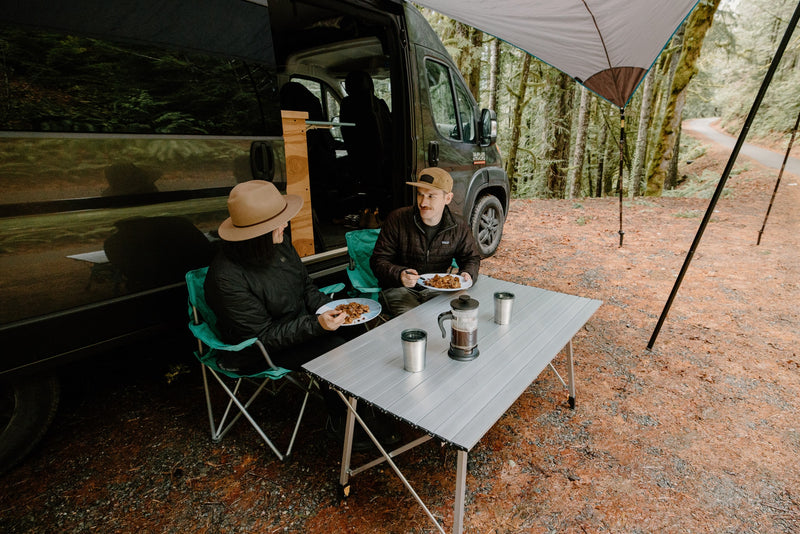 Two people enjoying an outdoor meal from their RV rental under an awning in a lush forest. Perfect for van life. — photo 14