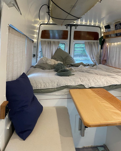Cozy RV rental interior features a comfortable bed, fold-down table, string lights, and overhead surfboard storage.