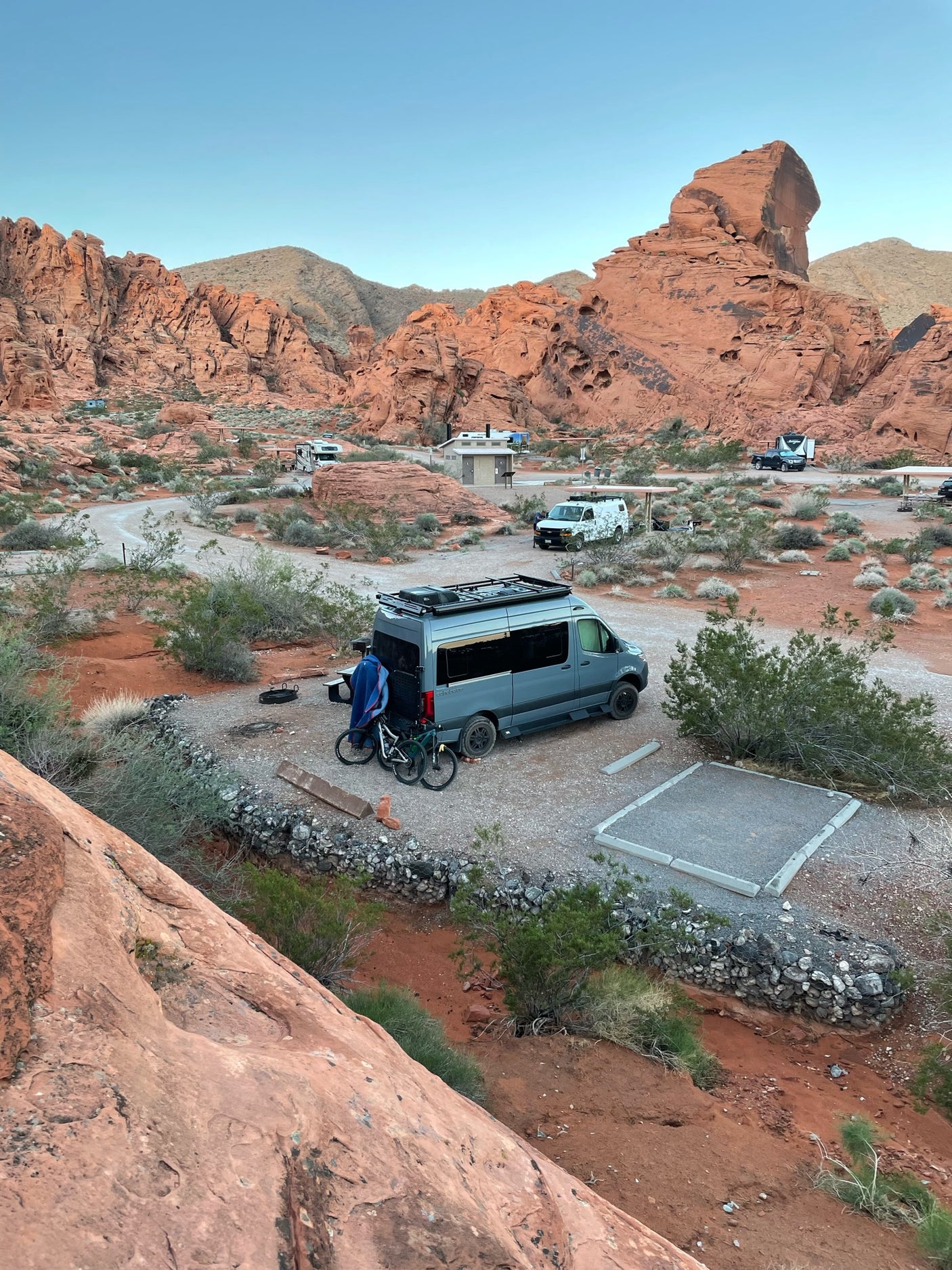 A gray camper van, a popular RV rental, is set up at a scenic desert campsite with red rocks and bikes.