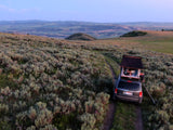 An SUV with a rooftop tent, a great RV rental option, parked on a dirt trail amidst sagebrush, offers serene camping at twilight.