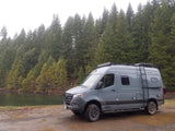 A gray Winnebago Revel 4x4 camper van RV rental parked by a forest lake on a muddy track. Ideal for off-grid adventures.