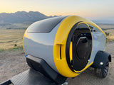Modern gray & yellow teardrop camper for RV rental, parked with mountains & sunset backdrop.