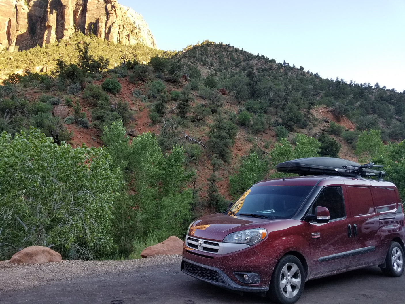 Red RV rental van with rooftop storage parked on a dirt road, facing a towering red rock canyon.