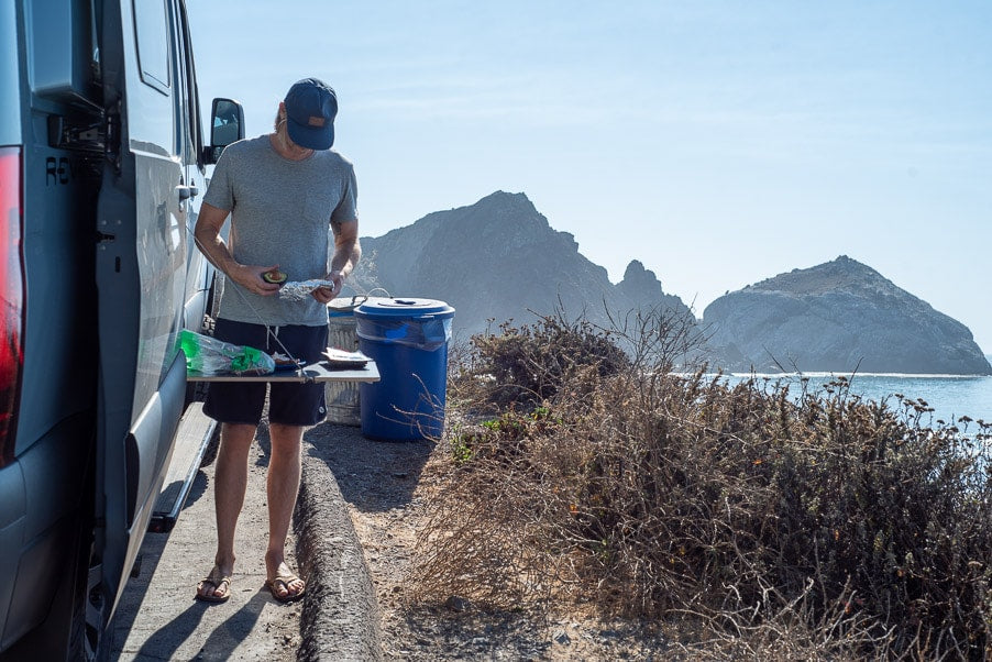 Man preps food by his RV rental, enjoying a scenic ocean view with rugged mountains. — photo 7