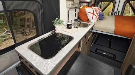 Cozy RV rental interior: kitchen with map-pattern counter, induction cooktop, sink, and comfy bed. Forest view through mesh window.