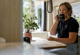 Enjoying coffee in a modern RV rental: A woman sips from a 'noovo' mug, with a plant and forest view.