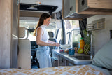 A woman prepares breakfast in a bright RV rental kitchen, with fresh fruit and a pastry on the counter. Ideal for mobile living.