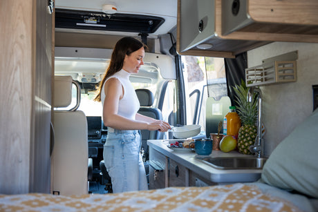 A woman prepares breakfast in a bright RV rental kitchen, with fresh fruit and a pastry on the counter. Ideal for mobile living.