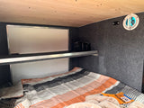 Cozy RV rental interior with a striped bed, overhead shelf holding a power station, and side wall vent for a comfortable sleep setup.