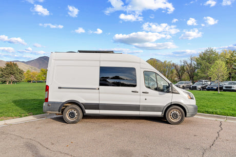 A white rv rental van, equipped with solar panels, parked on asphalt by a green park with mountains and a clear blue sky.