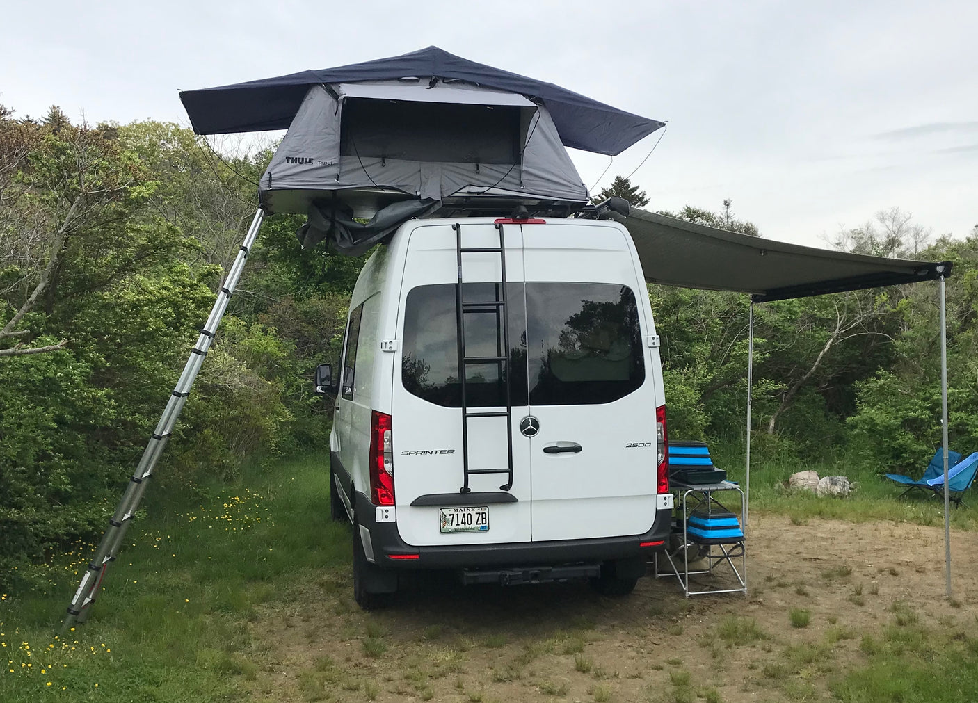 White RV rental Sprinter van with a rooftop tent and deployed awning, set up for outdoor camping.