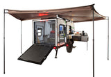 A compact off-road rv rental trailer, with a rear ramp, interior bed, and pull-out outdoor kitchen, under an awning.