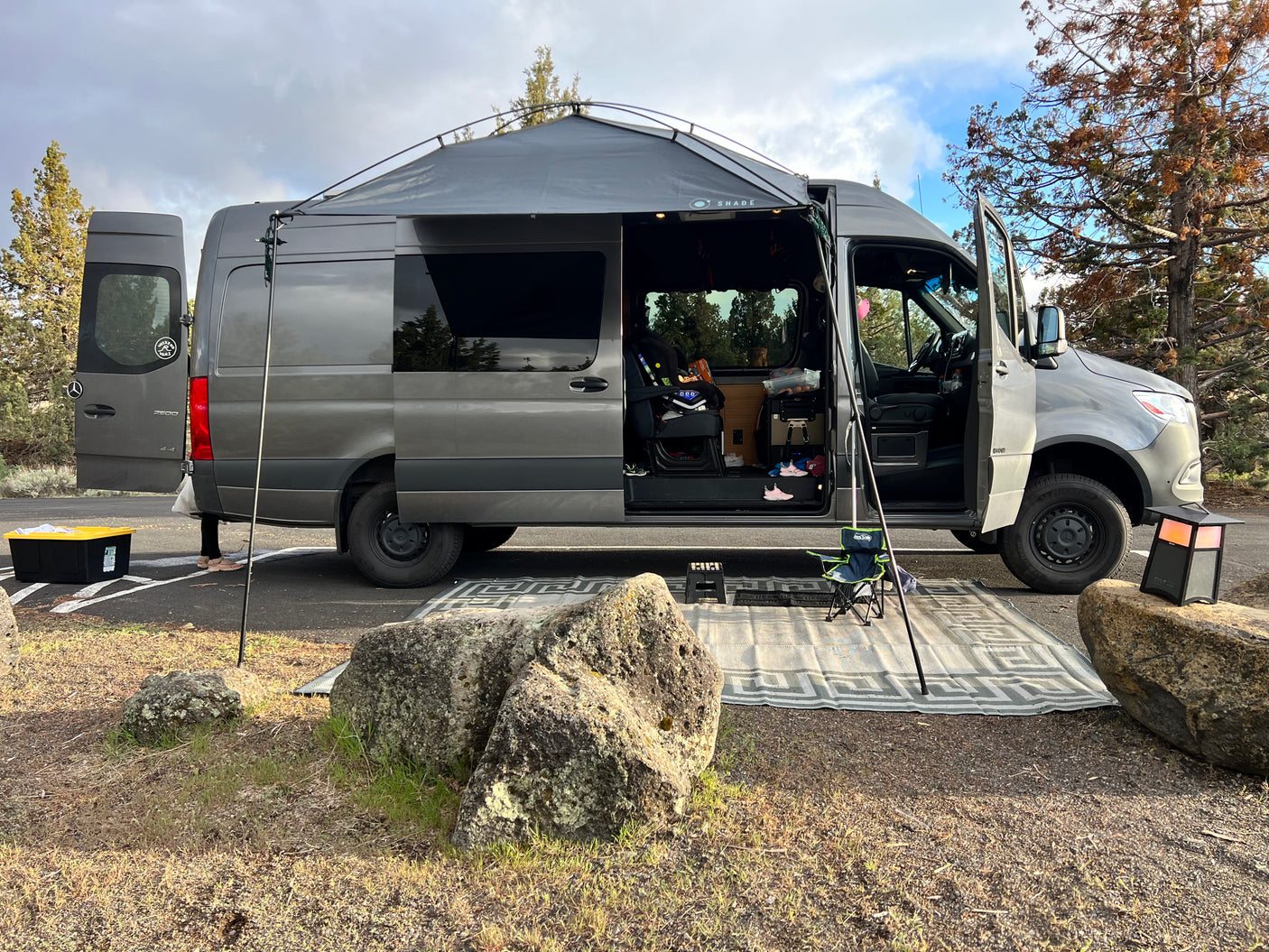 This grey RV rental van, with its large deployed awning and outdoor mat, provides a cozy campsite setup.