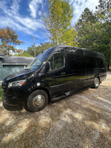 Black luxury RV rental van parked on a gravel driveway under a bright blue sky, amidst green trees.