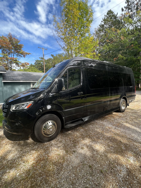 Black luxury RV rental van parked on a gravel driveway under a bright blue sky, amidst green trees.