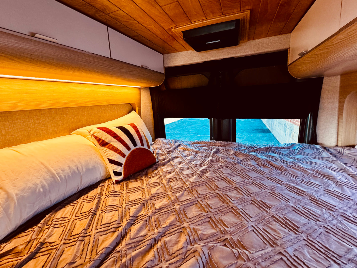 Comfortable RV rental bed featuring a textured blanket, decorative pillow, and warm overhead lighting with storage cabinets.
