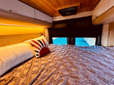 Comfortable RV rental bed featuring a textured blanket, decorative pillow, and warm overhead lighting with storage cabinets.
