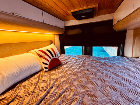 Comfortable RV rental bed featuring a textured blanket, decorative pillow, and warm overhead lighting with storage cabinets.