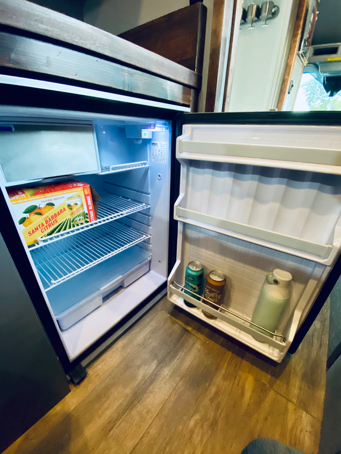 An RV rental's open mini-fridge, showing various beverages and a "Santa Barbara Citrus" box on shelves, set for travel.