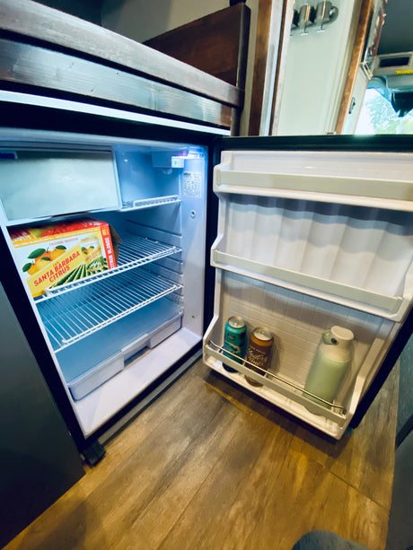 An RV rental's open mini-fridge, showing various beverages and a "Santa Barbara Citrus" box on shelves, set for travel.