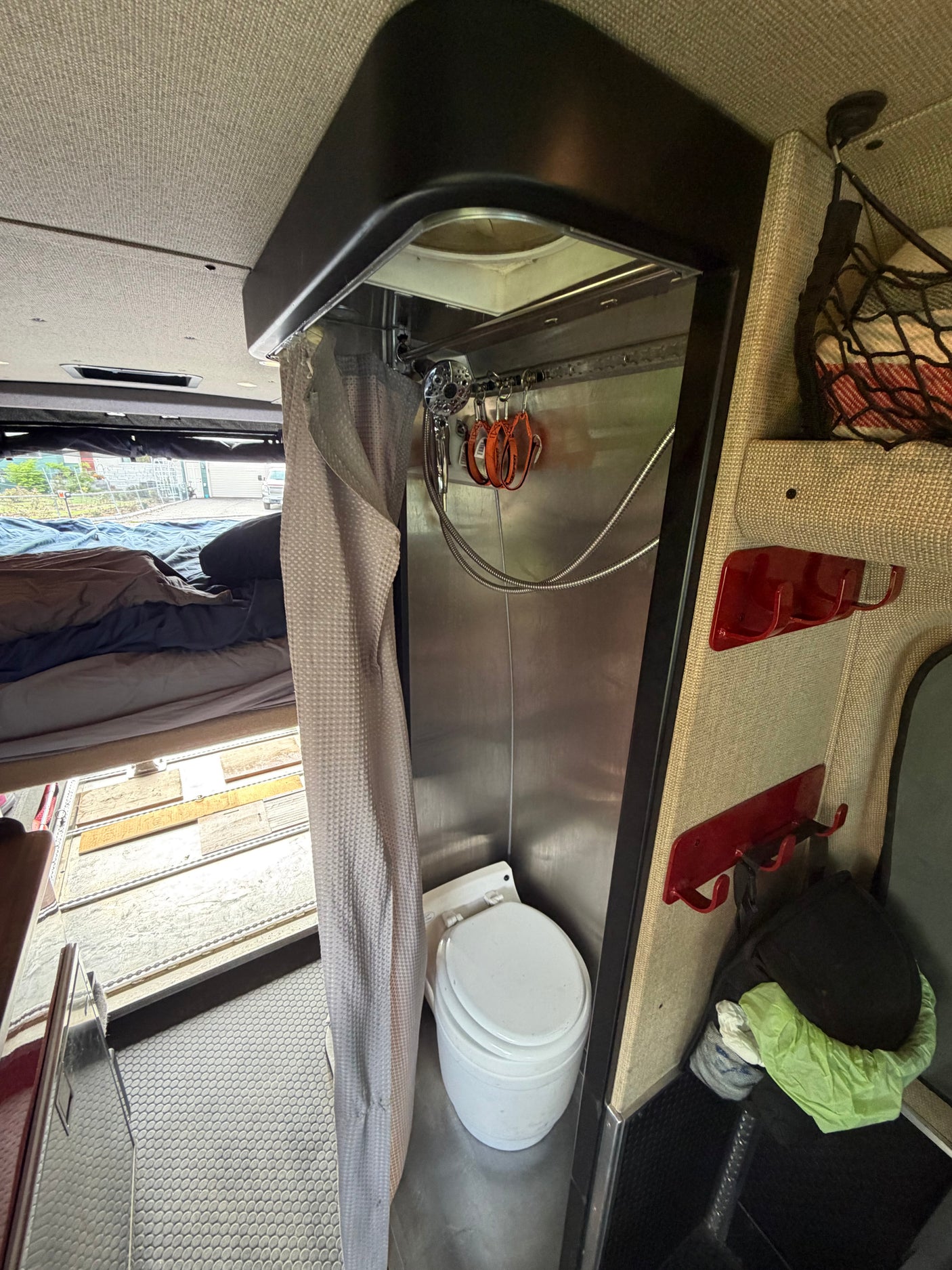 The interior of an RV rental showing a compact bathroom with a toilet and shower, next to an elevated bed. Textured walls and smart storage are visible.