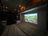 Inside an RV rental: a cozy bed faces a wall-mounted projector displaying a vibrant nature scene. Perfect for relaxing.