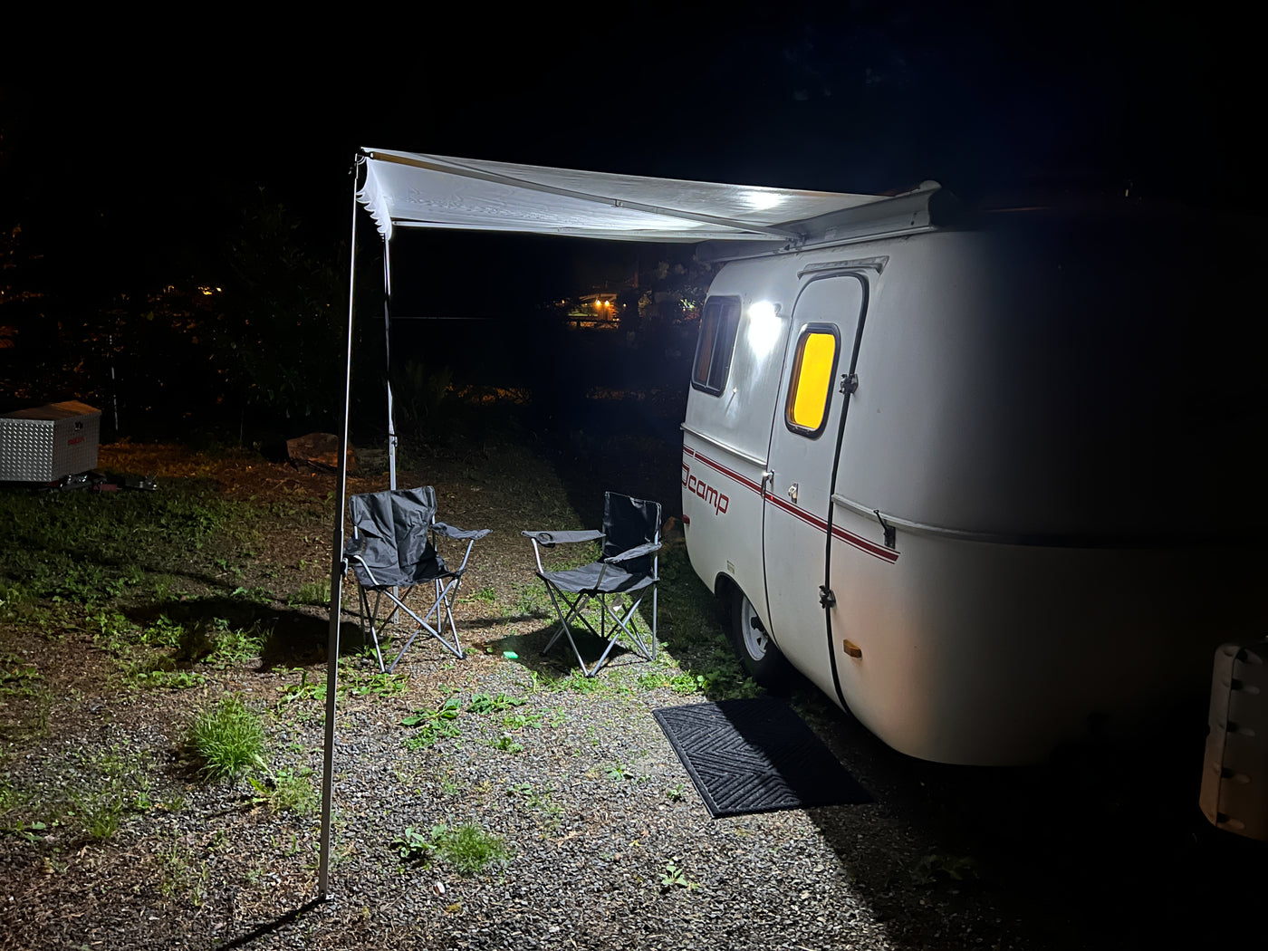 Cozy night at an RV rental campsite: small illuminated camper with awning extended, two chairs on gravel. — photo 16