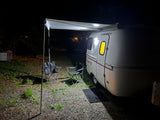Cozy night at an RV rental campsite: small illuminated camper with awning extended, two chairs on gravel.