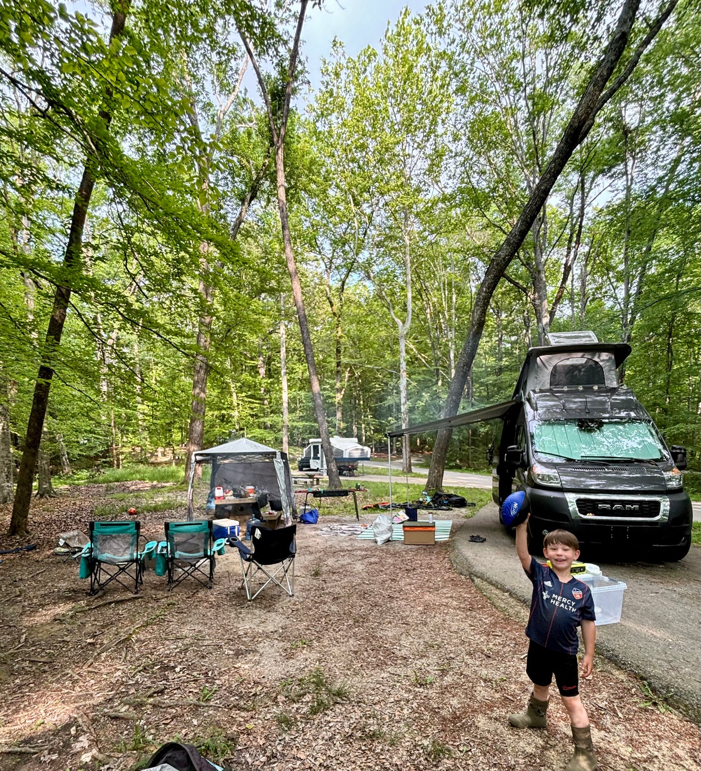 RV rental camping trip: A child poses happily at a shaded campsite with a black van, chairs, and tent setup.