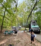 RV rental camping trip: A child poses happily at a shaded campsite with a black van, chairs, and tent setup.