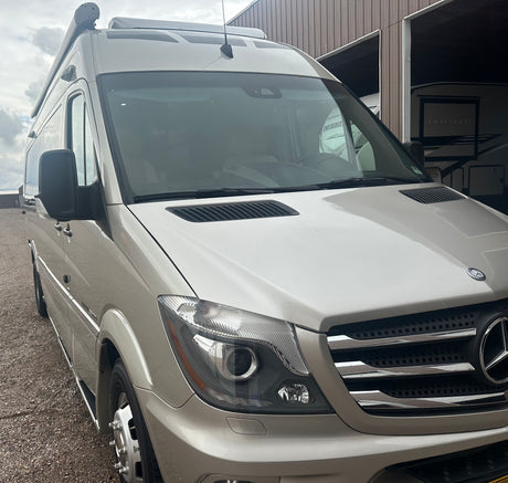 Tan Mercedes-Benz Sprinter RV rental, with a roof awning, parked on gravel next to a building.